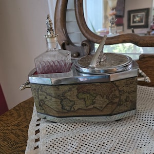May include: An antique vanity set featuring a decanter with a decorative stopper, a sundial, and a container with a world map design. The set is silver-toned with a wooden base and is displayed on a white lace doily.