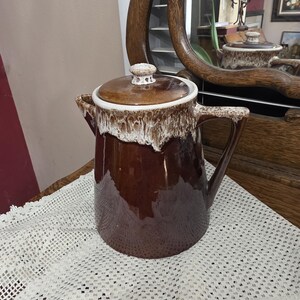May include: A brown ceramic coffee pot with a lid. The pot has a white and brown drip glaze around the top and handle. The handle is on the right side. The pot is sitting on a white lace tablecloth.