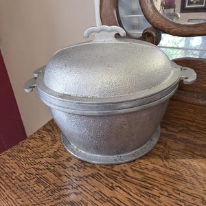 Puede incluir: Una cacerola vintage de metal plateado con una superficie texturizada. El plato tiene una tapa con un asa decorativa y asas laterales. Se asienta sobre una base redonda. Este artículo de cocina probablemente se utiliza para cocinar y servir.