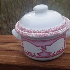 Puede incluir: Una pequeña olla de cerámica blanca con tapa, con un diseño rosa y blanco de dos cerdos enfrentados. La olla tiene un borde rosa y un diseño estampado rosa y blanco alrededor de la parte superior.
