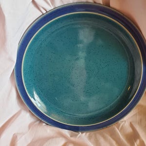 May include: A large, round, blue and green ceramic plate with a speckled glaze. The plate has a white rim and a blue band around the edge.
