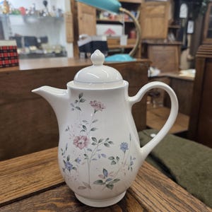 Puede incluir: Una tetera de cerámica blanca con tapa, con un diseño floral en tonos rosa, morado y azul. La tetera tiene un asa curva y un pico vertedor. Se encuentra sobre una superficie de madera.
