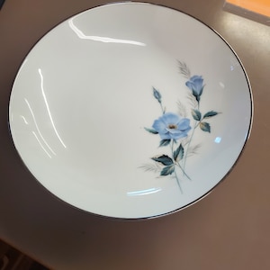 Puede incluir: Un tazón de cerámica blanca con un borde plateado y un diseño floral azul. El tazón tiene un diseño simple y elegante y es perfecto para servir una variedad de platos.