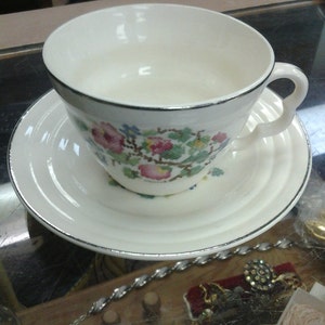 Puede incluir: Una taza de té y un platillo blancos con un patrón floral en tonos de rosa, verde y azul. La taza y el platillo tienen un borde plateado.