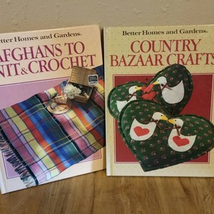Puede incluir: Dos libros de manualidades de Better Homes and Gardens. El primer libro se titula "Afghans to Knit & Crochet" y presenta una manta de cuadros de colores. El segundo libro se titula "Country Bazaar Crafts" y presenta una almohada en forma de corazón verde y roja con un par de pájaros blancos.