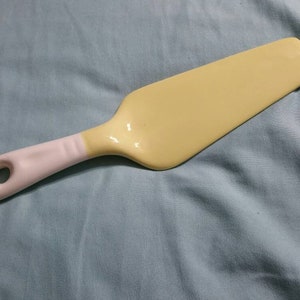May include: A yellow ceramic cake server with a white handle and a hole for hanging. The server has a flat, rectangular blade and a curved handle. The handle is approximately 4 inches long.