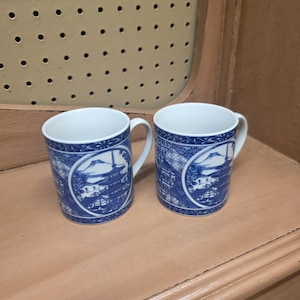 May include: Two white ceramic mugs with blue Japanese-style illustrations. Each mug features a mountain and pagoda scene within a circular frame, with additional blue patterns around the rim and handle.
