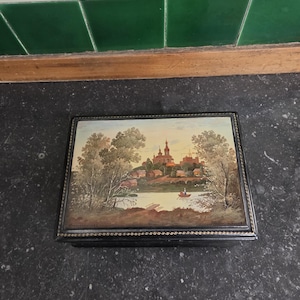May include: A rectangular black box with a painted landscape scene. The artwork depicts a river, trees, and buildings with red-orange roofs under a yellow sky. The box has a beaded border.