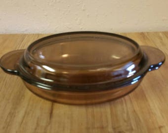 May include: A brown glass oval baking dish with a lid. The dish has two handles on the sides.