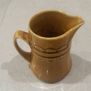 Puede incluir: Una pequeña jarra de cerámica de color marrón claro con asa y boquilla. La jarra tiene una banda decorativa con detalles festoneados alrededor del centro. El interior es de un tono marrón más oscuro.