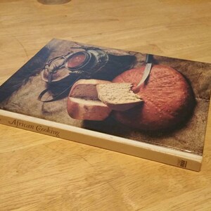 Könnte beinhalten: Ein gebundenes Buch mit dem Titel African Cooking, dessen Einband einen runden Laib Brot, ein Stück geschnittenes Brot und einen dunklen Metallkochtopf zeigt. Der Buchrücken ist hellbeige und der Titel ist in brauner Serifenschrift gedruckt.
