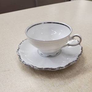 Könnte beinhalten: Ein weißes Tee-Set mit silbernem Rand. Die Tasse hat eine abgerundete Form und einen Henkel. Die Untertasse hat einen gewellten Rand.