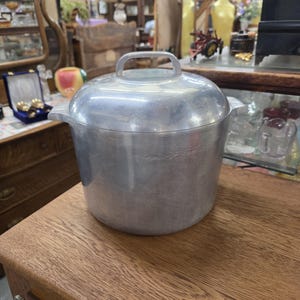 Könnte beinhalten: Ein Suppentopf aus gebürstetem Aluminium mit passendem Deckel. Der Topf hat einen zylindrischen Körper, zwei seitliche Griffe und einen Griff in der Mitte des Deckels. Die Metalloberfläche hat ein mattes Finish mit sichtbarer horizontaler Maserung.