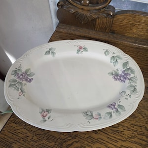May include: Oval serving platter with a white background and a decorative grape and leaf design in shades of purple, green, and pink. The platter has a scalloped edge and is placed on a wooden surface.