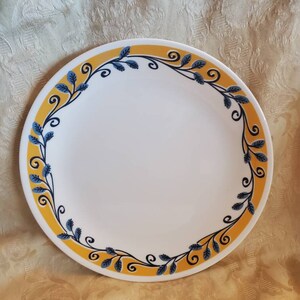 May include: A white ceramic dinner plate with a yellow and blue floral border. The border features a repeating pattern of stylised leaves and swirls.