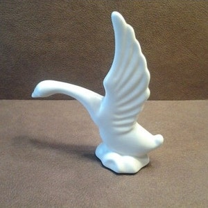 May include: A white ceramic figurine of a goose in flight. The goose has outstretched wings and a long, curved neck. The figurine is on a small, cloud-shaped base. The sculpture is a decorative item.