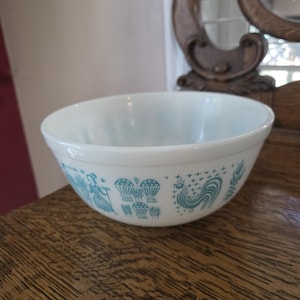 以下が含まれることがあります： 白のパイレックスボウル。ターコイズブルーの模様で、様式化された人物と雄鶏が描かれています。ボウルは丸みを帯びた形状で、滑らかで光沢のある仕上がりです。ヴィンテージのキッチン用品です。