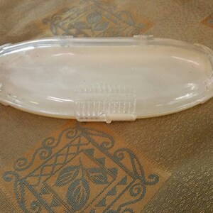 May include: A clear plastic oval-shaped container with a textured lid. The container is empty and sitting on a patterned fabric surface.