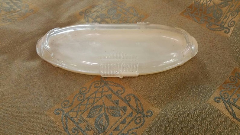 May include: A clear plastic oval-shaped container with a textured lid. The container is empty and sitting on a patterned fabric surface.
