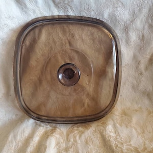 以下が含まれることがあります： 透明な中央と上部に小さな丸いノブが付いた正方形の茶色のガラスの蓋