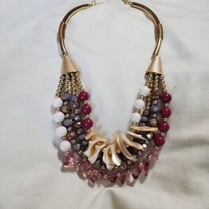 May include: A multi-strand necklace with gold metal accents and a variety of beads, including white, pink, purple, and red. The necklace features a unique design with shell-shaped beads and a gold metal clasp.