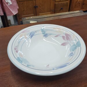 May include: A white ceramic serving bowl with a floral design in pastel colors. The bowl has a wide, shallow shape with a light blue rim and decorative bands. The floral pattern includes pink, blue, and yellow leaves.