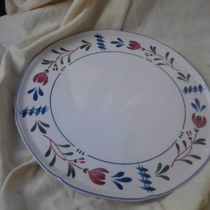 Puede incluir: Plato de cerámica blanca con borde festoneado, adornado con un motivo floral. El diseño presenta detalles en rojo, azul y verde, con un borde azul. Este plato decorativo es adecuado para servir alimentos.