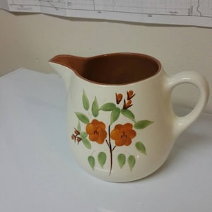 Puede incluir: Una jarra de cerámica blanca con un borde y un asa marrones. La jarra tiene un diseño floral con flores naranjas y hojas verdes.