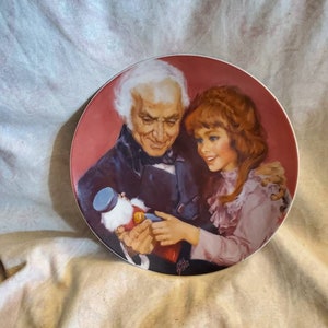 May include: A vintage porcelain plate with a red background featuring a painting of an older man with white hair and a girl with red hair. The man is holding a nutcracker in his hand and the girl is looking at the nutcracker. The plate has a gold rim.