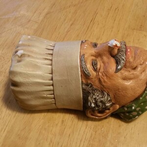 May include: A ceramic wall decoration of a chef's head wearing a white chef's hat with a green and white polka dot bandana and a red bow. The chef has a white moustache and a white beard.