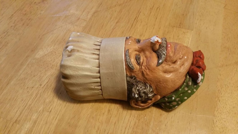 May include: A ceramic wall decoration of a chef's head wearing a white chef's hat with a green and white polka dot bandana and a red bow. The chef has a white moustache and a white beard.
