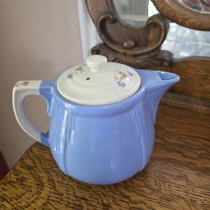 May include: A light blue ceramic teapot with a white lid and handle. The lid is decorated with small floral patterns in shades of pink, blue, and yellow. The teapot is sitting on a wooden surface.