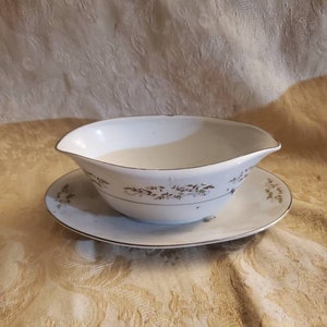 May include: White porcelain gravy boat with a gold trim and floral design. The boat is sitting on a matching saucer.