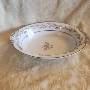 May include: White oval serving dish with silver trim and a silver floral design. The dish has two handles on the sides.