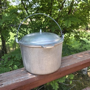May include: A silver-colored aluminum pot with a textured exterior finish, featuring a matching lid with a small central knob and a sturdy wire bail handle attached to two side loops, designed for cooking or storage purposes.