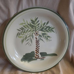 May include: A white ceramic dinner plate with a green rim and a hand-painted design of a palm tree with green leaves and a brown trunk.