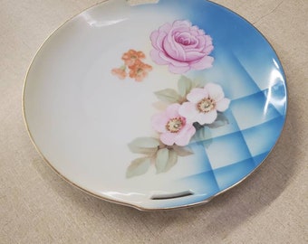 May include: A white porcelain serving plate with a blue and white chequered pattern and a floral design featuring pink and orange roses. The plate has two gold handles on the sides.