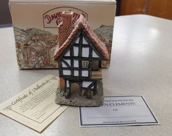 May include: A small, detailed, hand-painted ceramic cottage with a brown roof, white walls, and black trim. The cottage has a chimney and a small door. The cottage is sitting on a white base. The text on the box reads "David Winter Cottages".