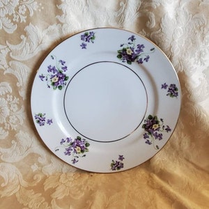 May include: A white ceramic dinner plate with a gold rim and a floral design featuring purple and yellow violets.