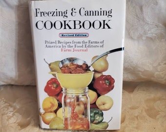May include: A white cookbook with the title "Freezing & Canning Cookbook" in red and black text. The subtitle is "Revised Edition" in a smaller font. The cover features a yellow funnel pouring red sauce into a glass jar. There are various fruits and vegetables surrounding the jar, including apples, pears, peppers, and tomatoes.