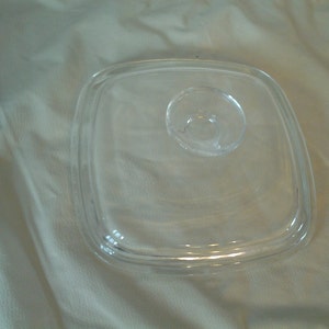 May include: A clear, square glass lid with a rounded handle. The lid has a slightly raised edge and is designed to fit a square-shaped dish. The glass is transparent, allowing visibility of the contents below.