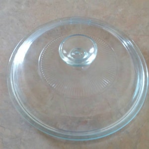 May include: A clear glass lid with a knob on top. The lid has a textured pattern on the bottom.