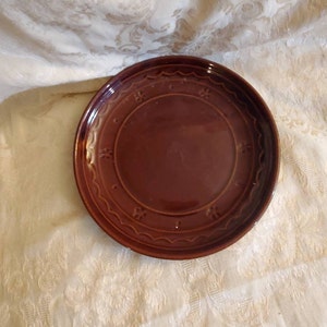 Könnte beinhalten: Ein brauner Keramikteller mit einem gewellten Rand und einem dekorativen Muster um den Rand. Der Teller ist leer.