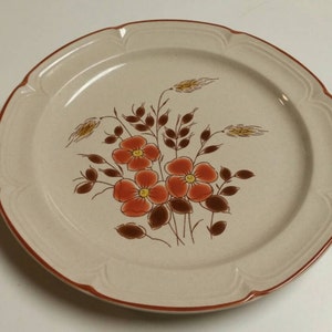 May include: A beige ceramic dinner plate with a brown rim and a floral design featuring orange and brown flowers and wheat stalks.