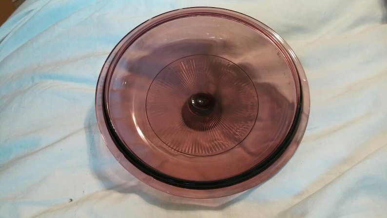 May include: A clear glass lid with a purple tint and a raised center knob. The lid has a decorative pattern of concentric circles.