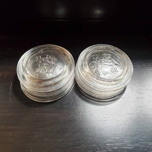 Può includere: Una pila di 10 coperchi per barattoli di vetro trasparente con un design in rilievo e il testo "Ball" e "Wide Mouth" sul coperchio superiore.
