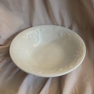 May include: A white, round bowl with a decorative embossed fruit and leaf pattern around the rim. The bowl has a smooth, glossy finish and is set against a neutral background.