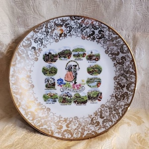 May include: A white ceramic plate with a gold rim and a floral design. The center of the plate features a map of Austria with illustrations of different cities and landmarks. The text "Grüß aus Österreich" is written in the center of the map.