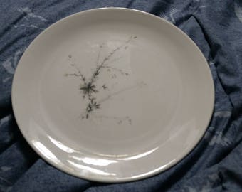 May include: A white ceramic plate with a delicate floral design in shades of gray and light pink. The design features a stylized branch with small flowers and leaves.
