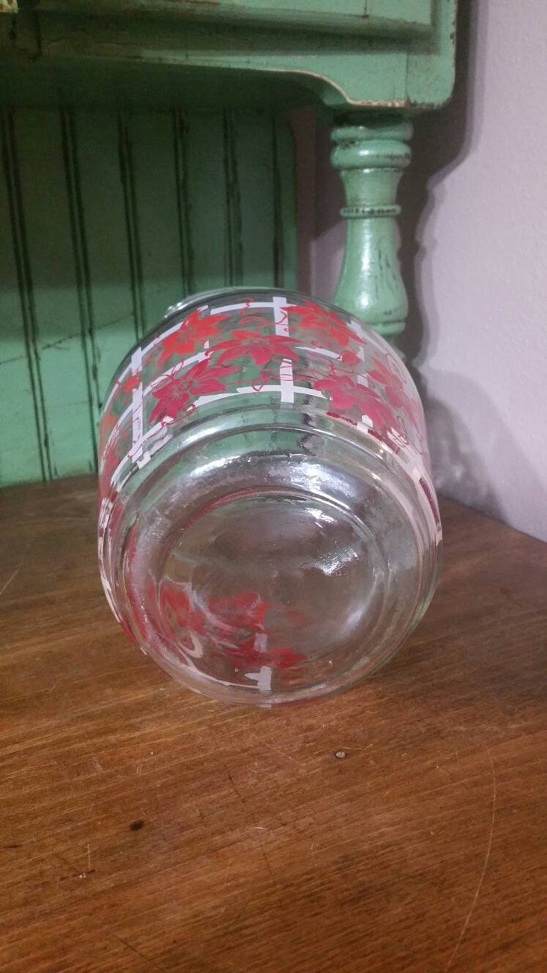 May include: A clear glass jar with a red and white floral pattern. The jar has a wide mouth and a flat bottom.